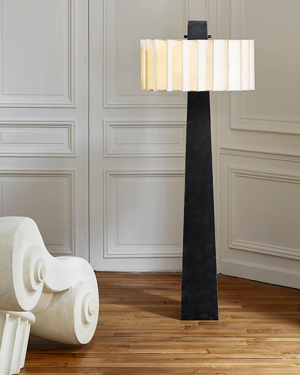 Lámpara de pie columna con pantalla plisada Vestala – junto a una butaca blanca escultórica