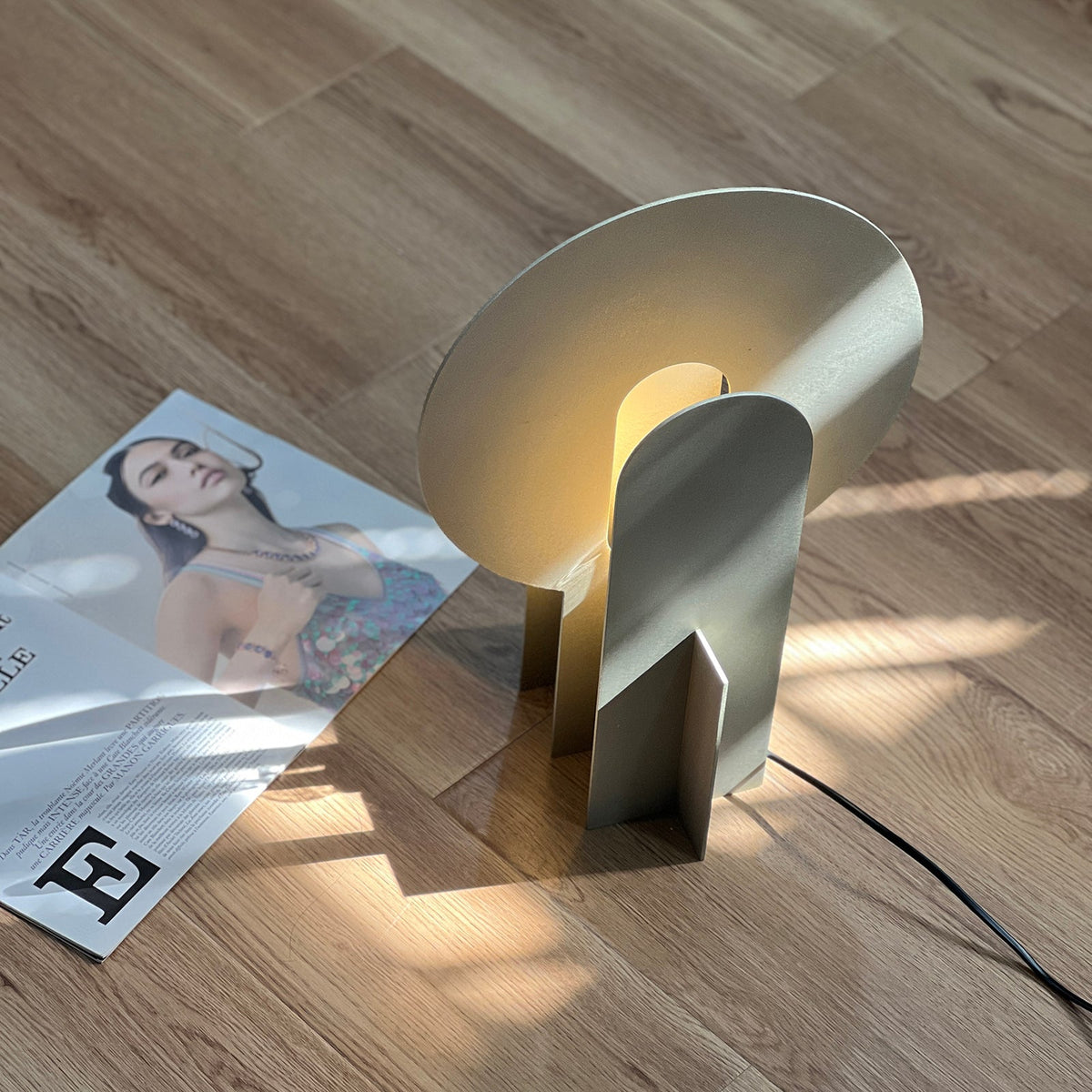 Lámpara de mesa de acero con arco Mardela – encendida en el suelo junto a una revista