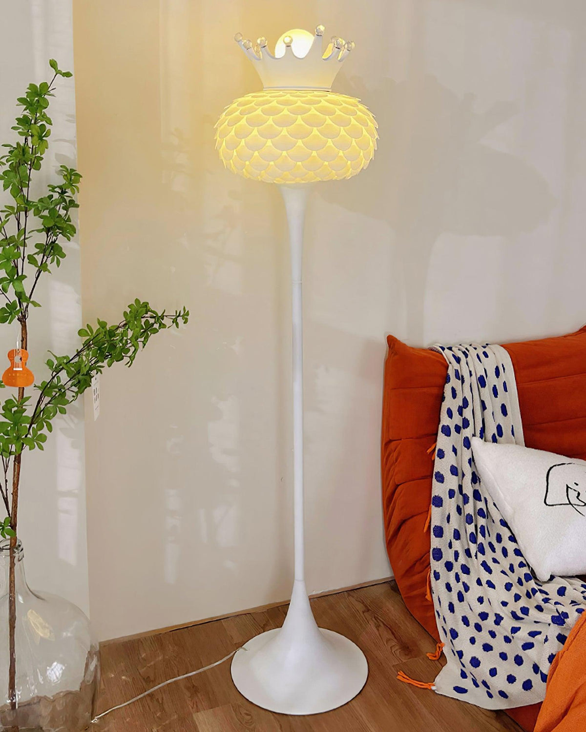 Lámpara de pie blanca con corona y pantalla texturizada Halvira – encendida junto a un cojín naranja
