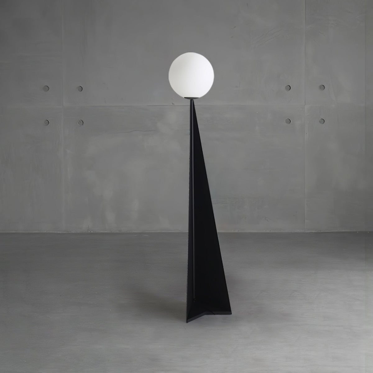 Lámpara de pie con esfera blanca y base triangular negra Pelia – frente a una pared de hormigón