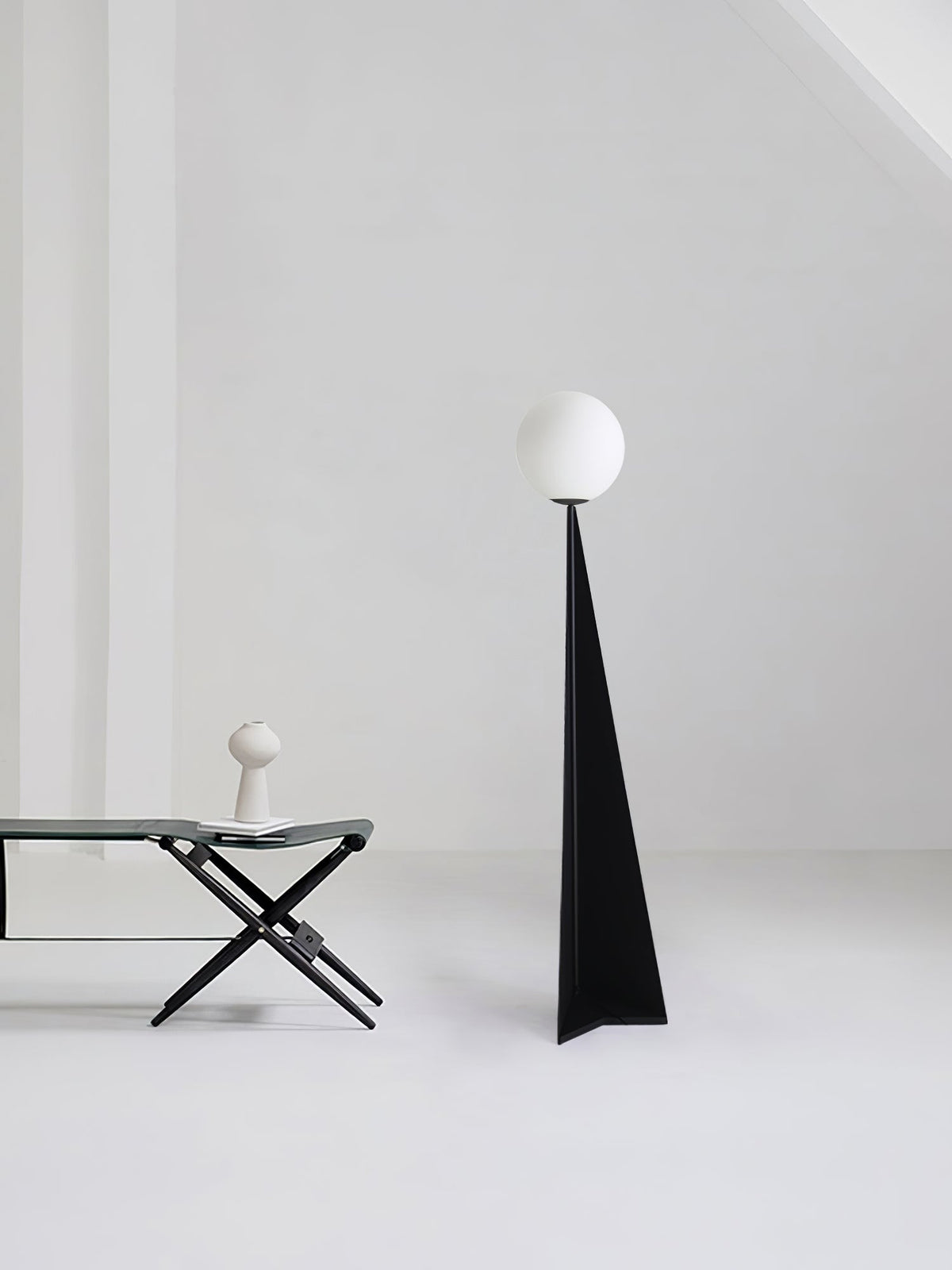 Lámpara de pie con esfera blanca y base triangular negra Pelia – junto a una mesa auxiliar plegable