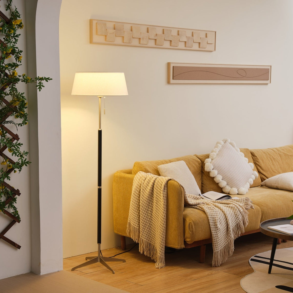 Lámpara de pie con base trípode y pantalla de tela blanca Iluma – encendida en un rincón junto a un sofá