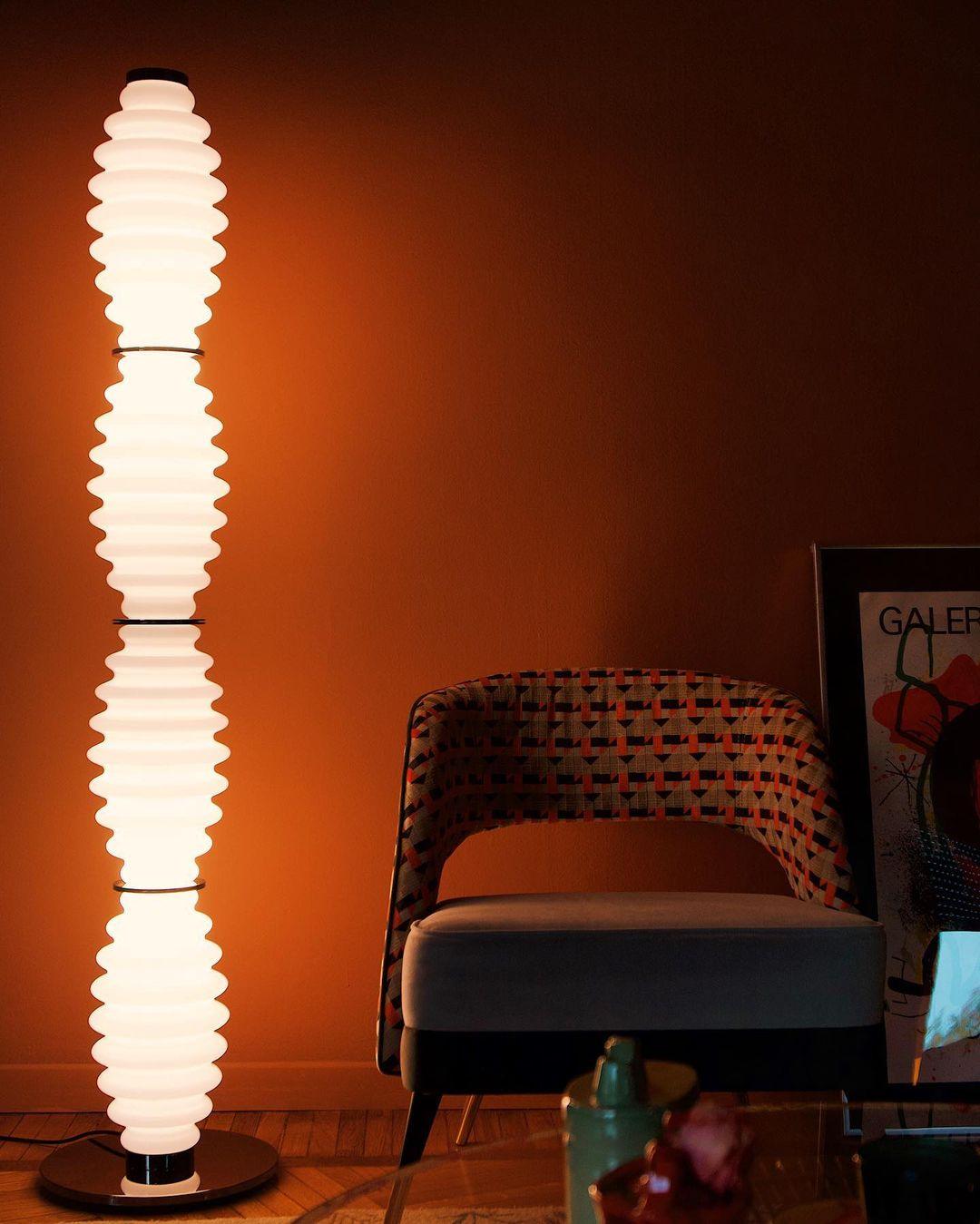 Lámpara de pie de cristal Tulara – junto a una butaca bajo una luz anaranjada