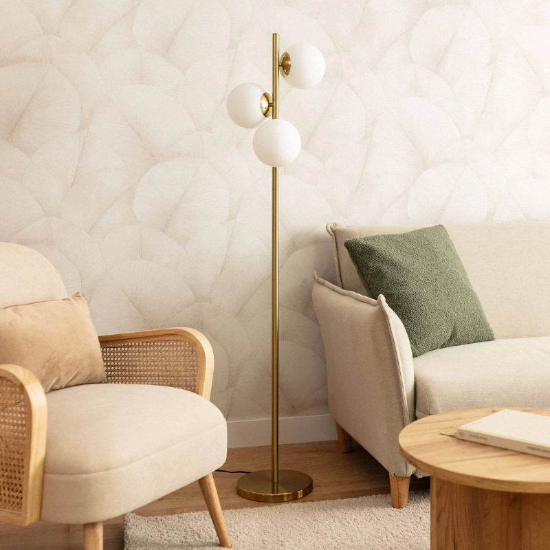 Lámpara de pie dorada Tadela con tres globos de cristal – apagada junto a un sillón