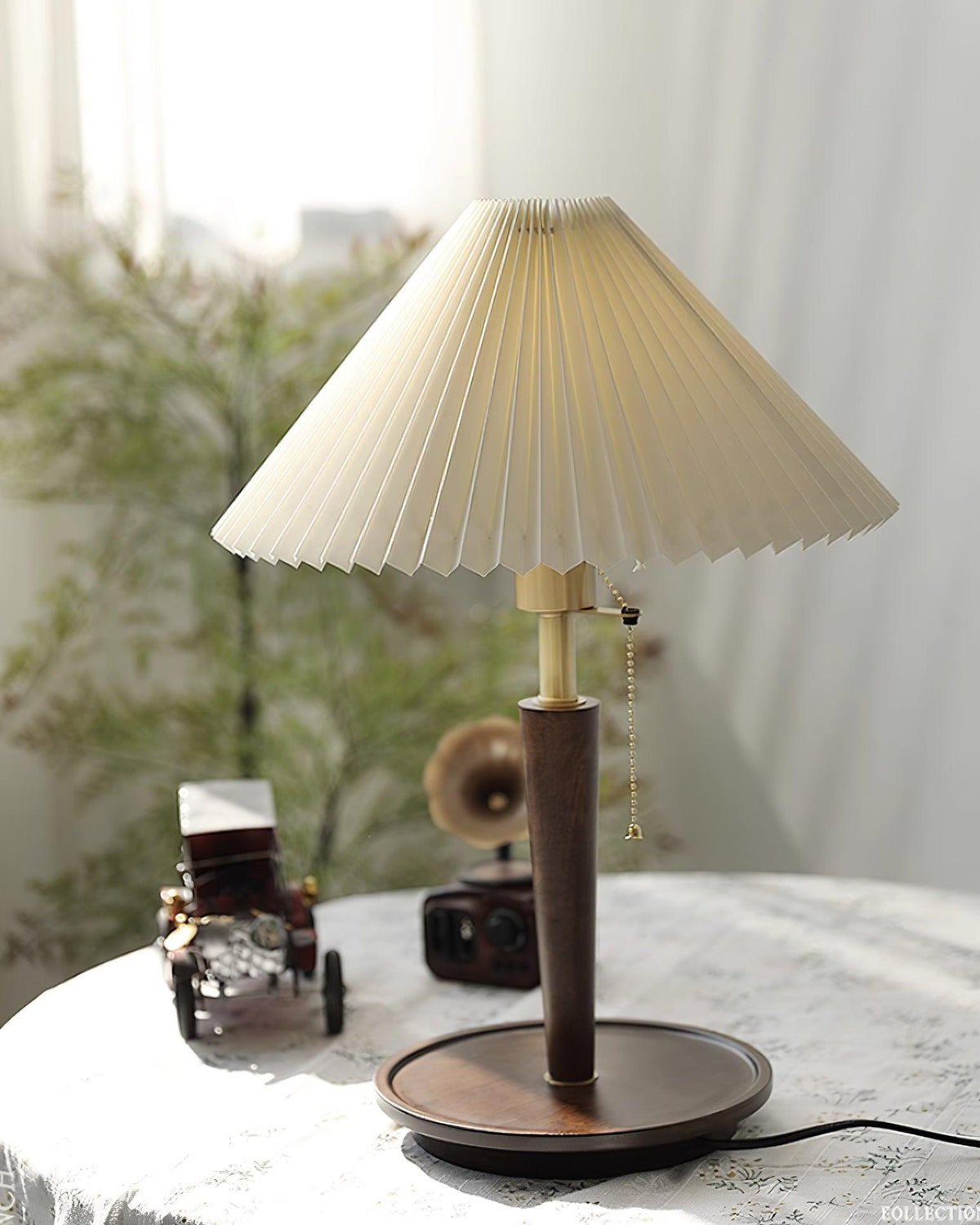 Lámpara de mesa plisada Ripela – sobre una mesa con figuras decorativas