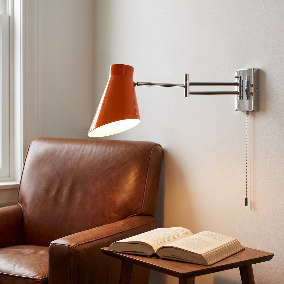Aplique de pared con brazo extensible Kurela – pantalla naranja junto a un sillón de cuero