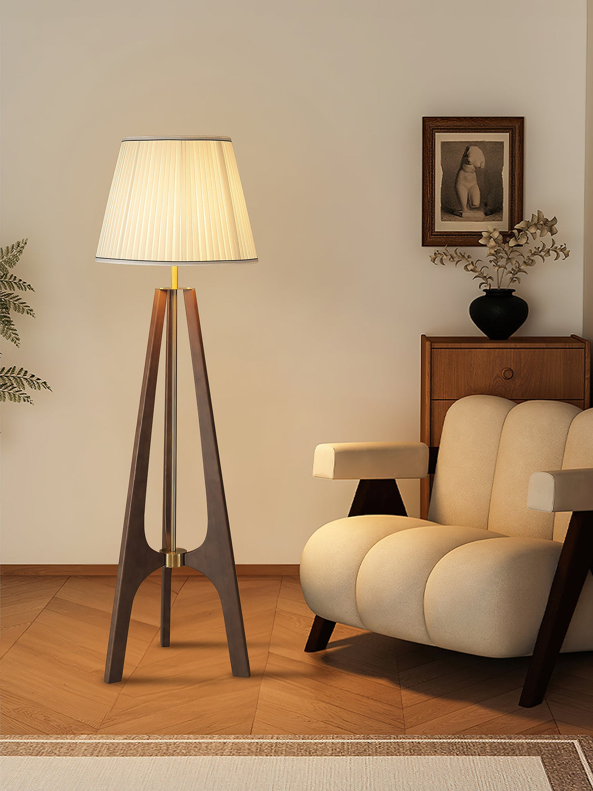 Lámpara de pie trípode de madera con pantalla plisada blanca Sabina – junto a un sillón de líneas redondeadas