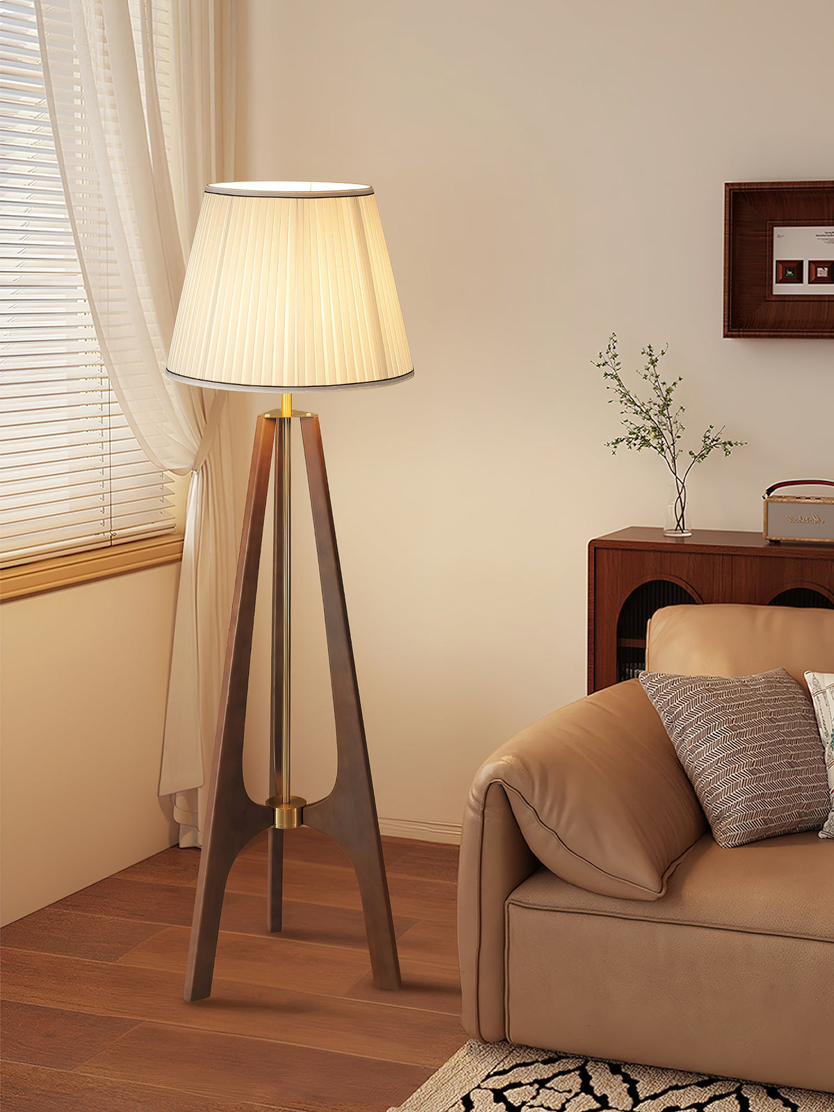 Lámpara de pie trípode de madera con pantalla plisada blanca Sabina – junto a un sofá de cuero y una cortina