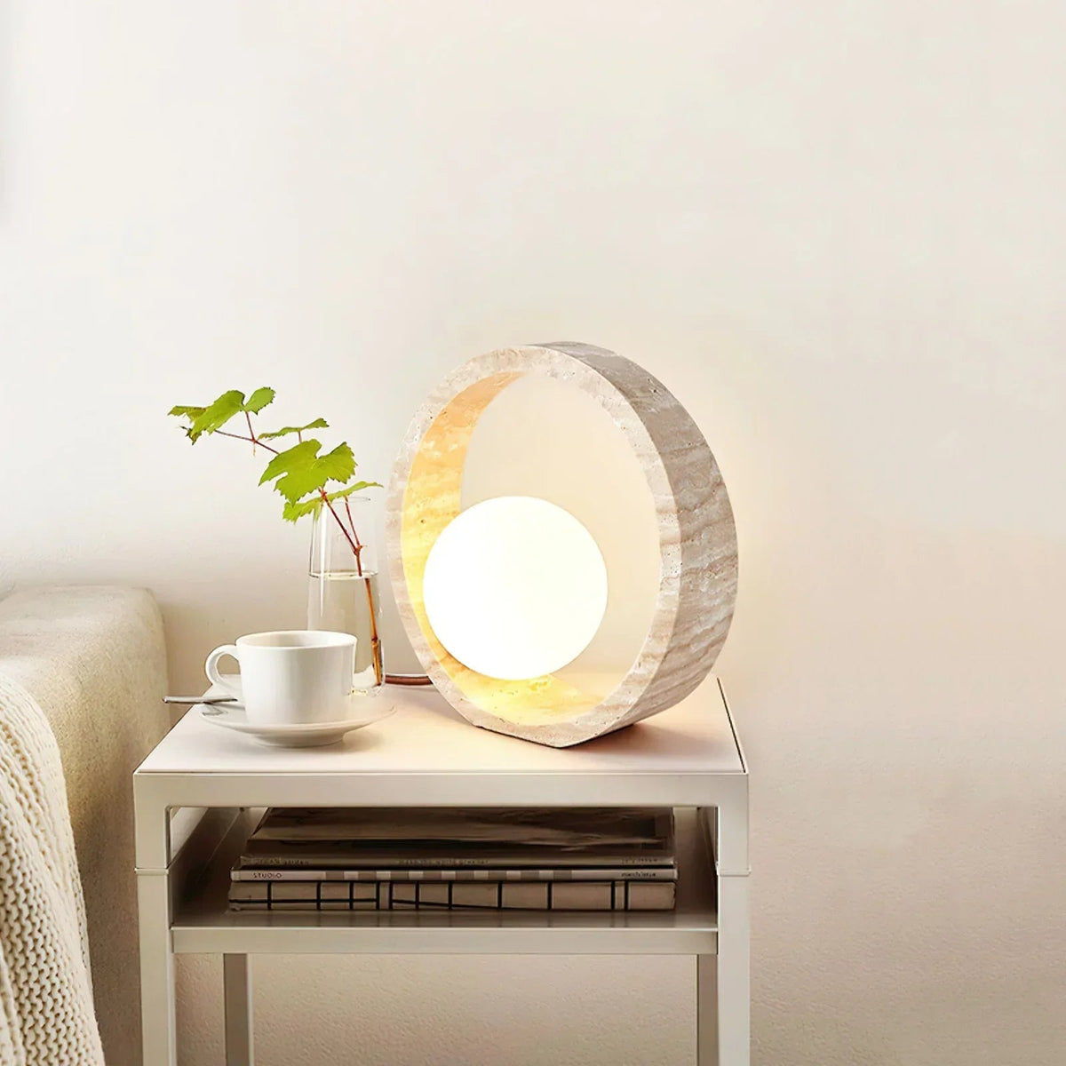 Lámpara de mesa de travertino Anella – encendida sobre una mesita blanca junto a una planta