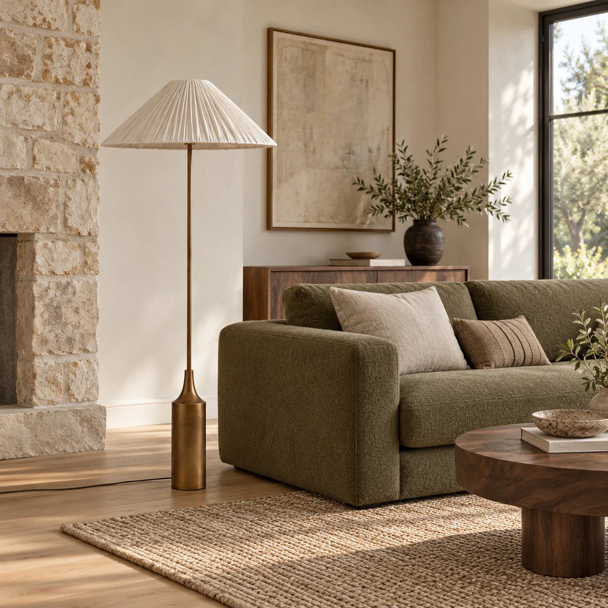 Lámpara de pie con pantalla plisada de lino y base latón Soleira – junto a un sofá verde en el salón