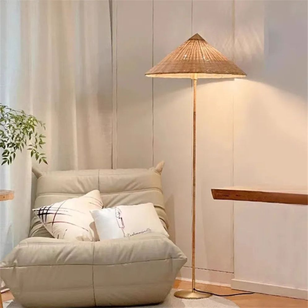 Lámpara de pie con pantalla cónica Bacora – de ratán encendida junto a un sillón