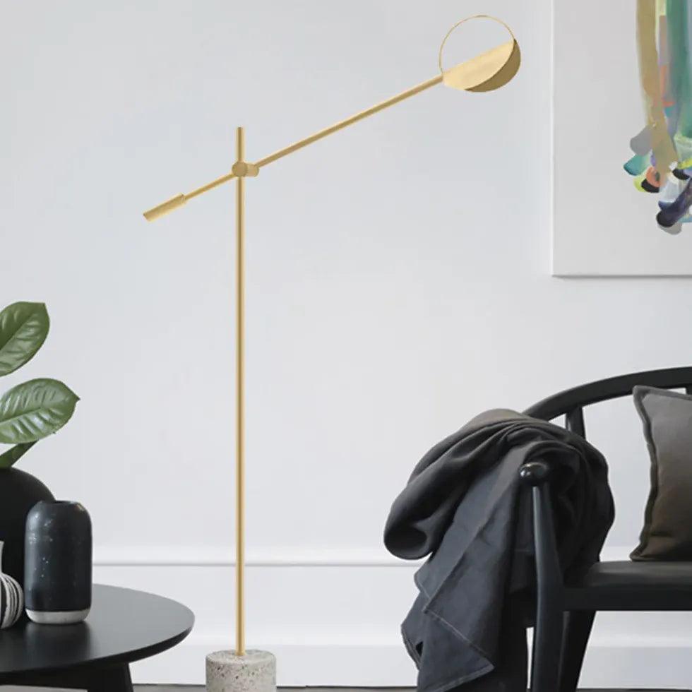Lámpara de pie con brazo basculante y base de terrazo Dorvela – dorada junto a una silla