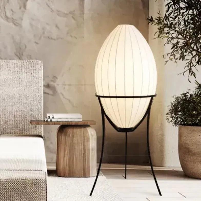 Lámpara de pie ovalada de tela Ilvia – junto a una mesa de madera y plantas en un salón de tonos naturales