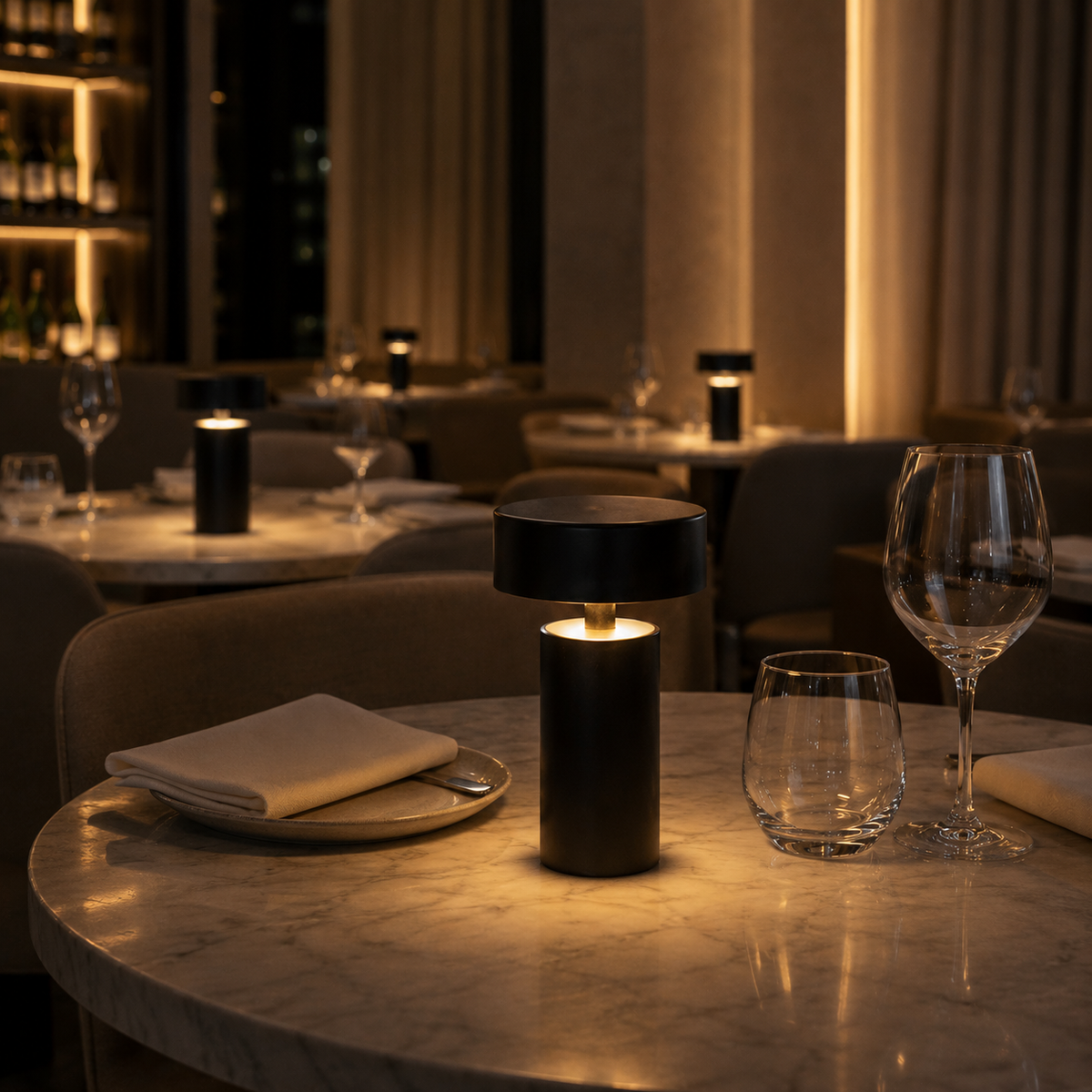 Lámpara de mesa LED recargable Kirena – negra encendida en una mesa de restaurante