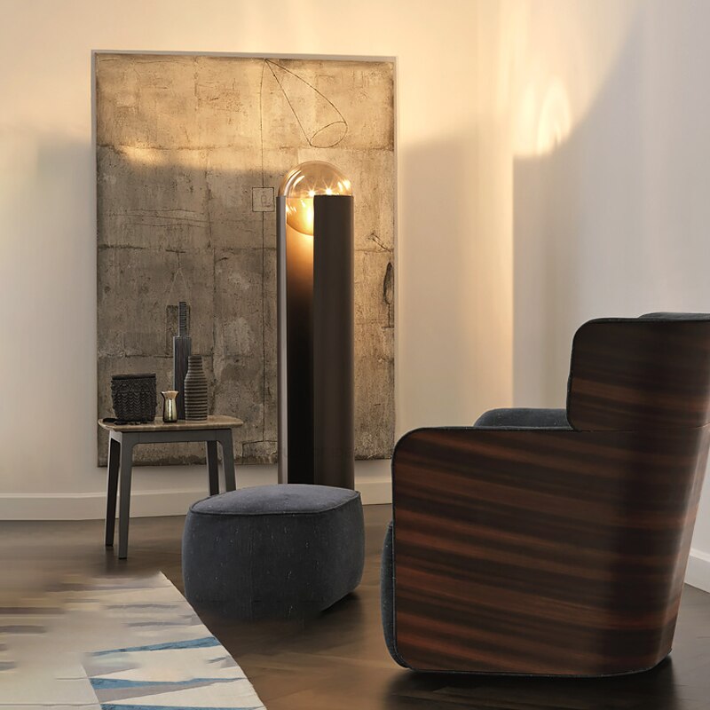 Lámpara de pie columna Velatra – junto a un sillón de madera