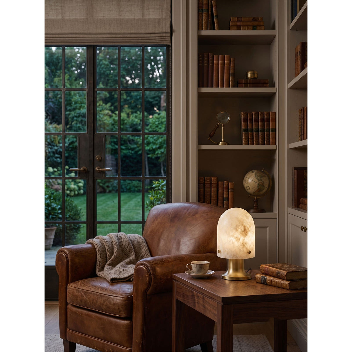 Lámpara de mesa de alabastro Quarzela – encendida en una biblioteca junto a un sillón de cuero