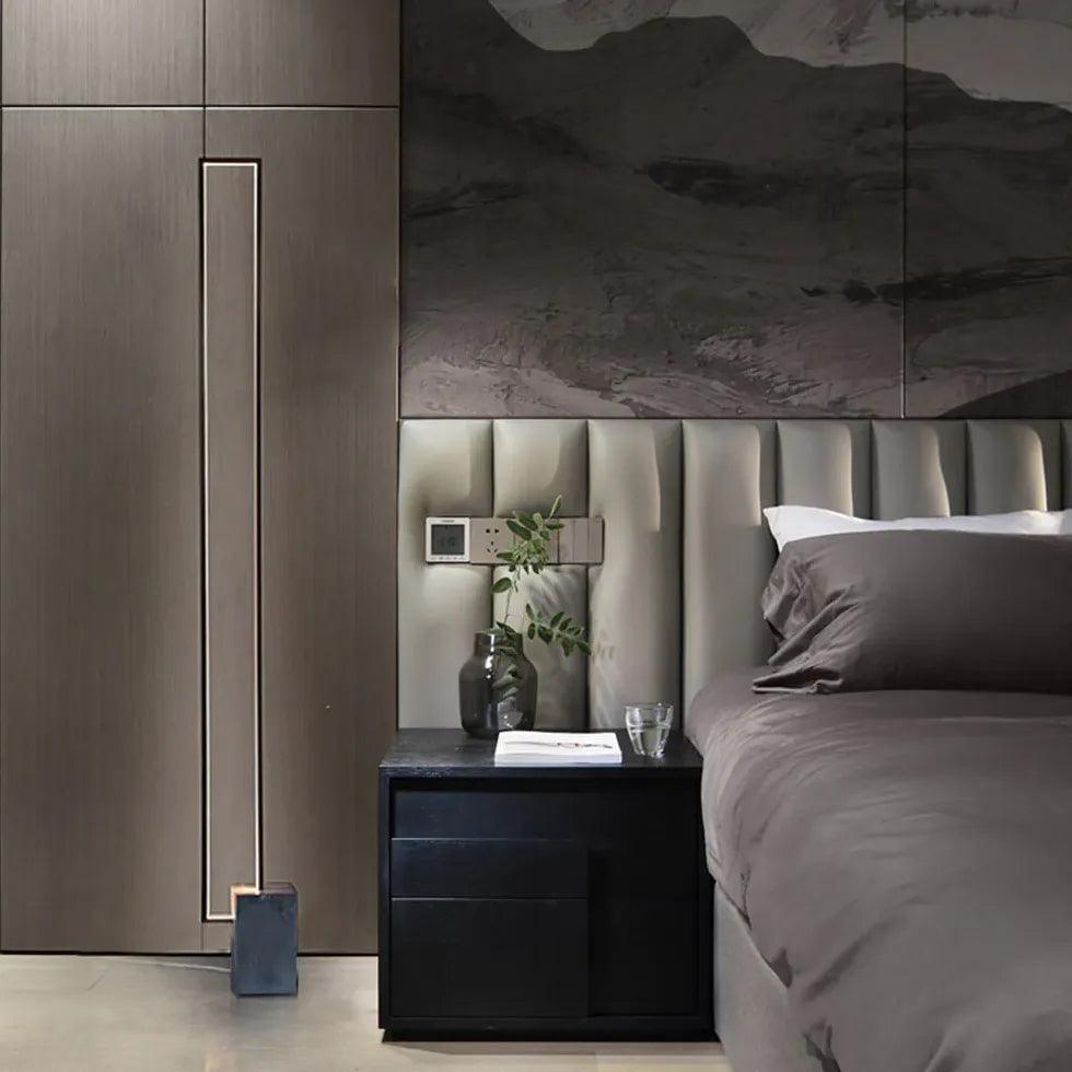 Lámpara de pie LED rectangular minimalista de metal negro Riela – junto a una mesilla en un dormitorio