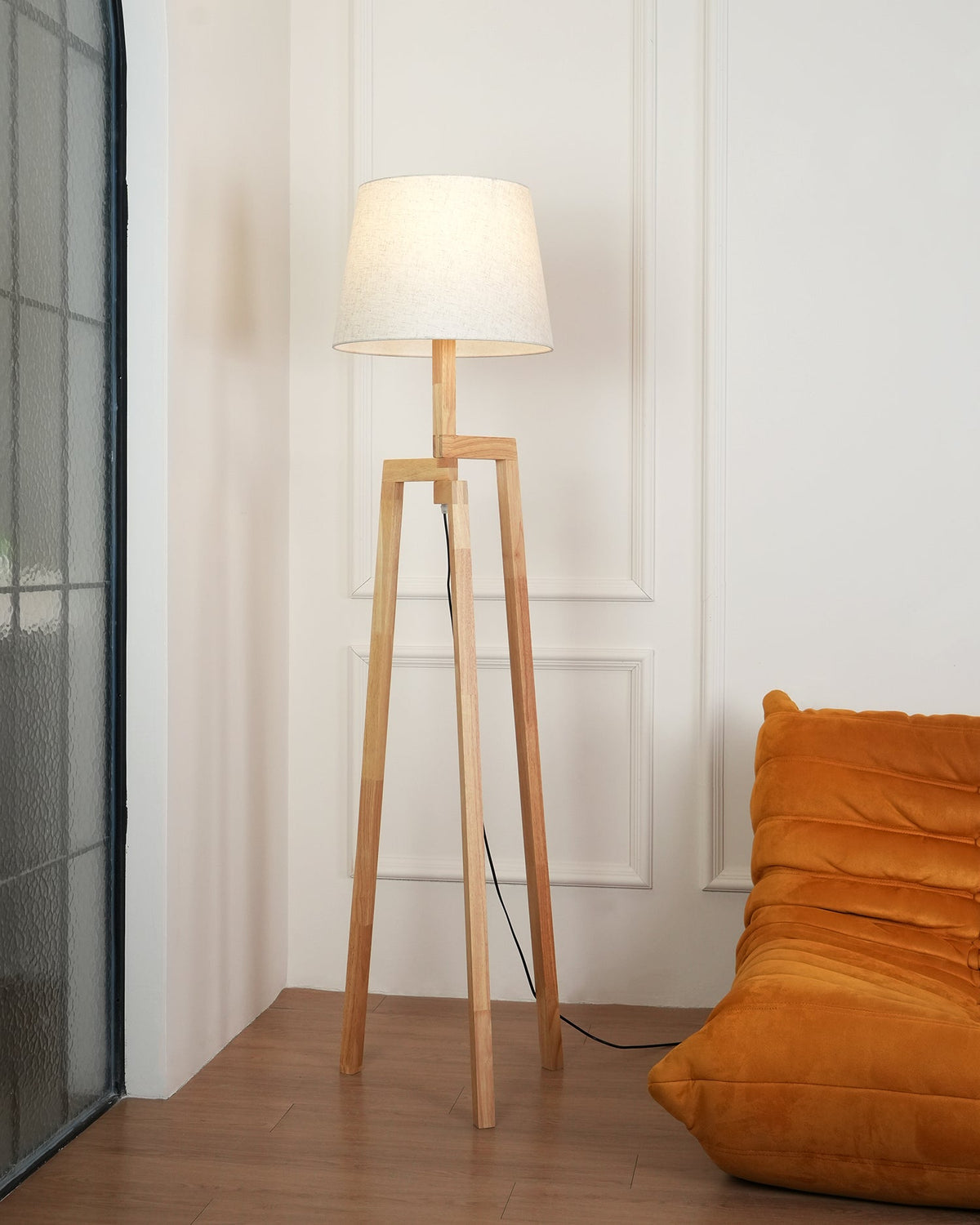 Lámpara de pie trípode de madera Tresalba – encendida junto a un sillón naranja
